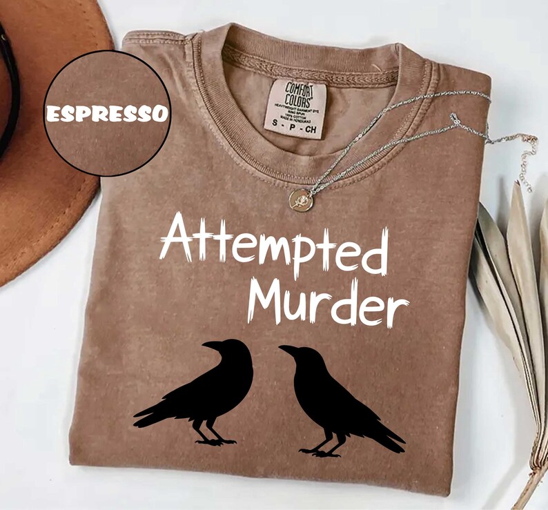May include: Espresso-colored t-shirt with the words "Attempted Murder" in white, along with two black crow silhouettes. The shirt has a crew neck and short sleeves. A silver necklace with a pendant is draped across the shirt.