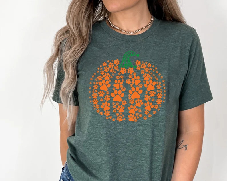 Peut inclure: T-shirt vert fonc&eacute; avec un motif de citrouille orange compos&eacute; d'empreintes de pattes. La citrouille a une tige verte. Le t-shirt est de style col rond et est fait d'une mati&egrave;re douce. Id&eacute;al pour l'automne.