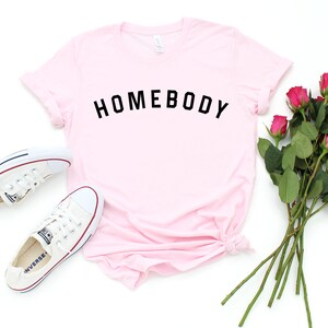 Puede incluir: Una camiseta rosa con el texto "HOMEBODY" impreso en negro.