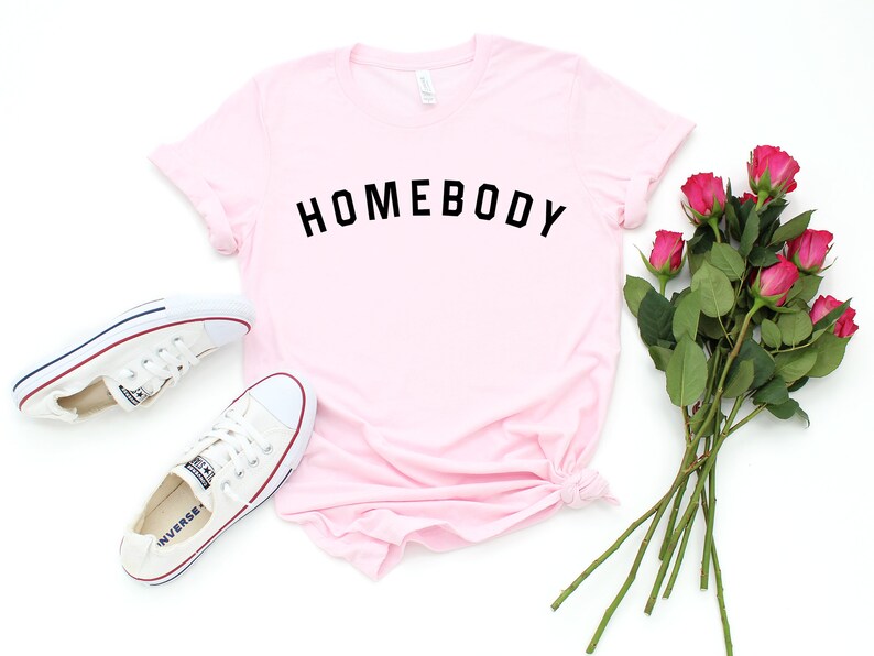 Puede incluir: Una camiseta rosa con el texto "HOMEBODY" impreso en negro.