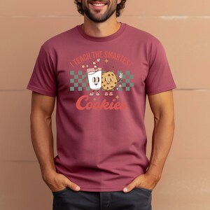 May include: A maroon t-shirt with a cartoon illustration of a glass of milk and a cookie. The text on the shirt reads "I teach the smartest cookies".