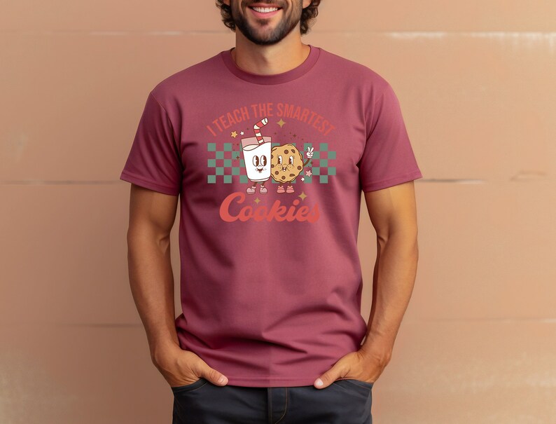 May include: A maroon t-shirt with a cartoon illustration of a glass of milk and a cookie. The text on the shirt reads "I teach the smartest cookies".