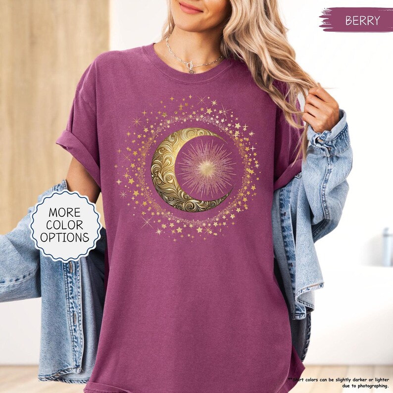 May include: Berry-colored t-shirt featuring a gold crescent moon and starburst design, surrounded by gold stars. The shirt has rolled-up sleeves and a round neckline. The words "MORE COLOR OPTIONS" are in a white bubble on the left side of the shirt.