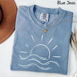 Puede incluir: Una camiseta azul vaquero con un gr&aacute;fico blanco de sol y olas. La camiseta tiene cuello redondo y mangas cortas. Un collar dorado con un colgante est&aacute; drapeado sobre la camiseta.