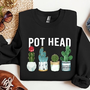 May include: Black sweatshirt with the text "POT HEAD" in white lettering. The sweatshirt features a graphic of four potted cacti with different colored flowers and patterns.