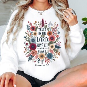 Peut inclure: Sweat-shirt blanc orn&eacute; d'un motif floral avec le texte "Trust in the Lord with all your heart" et "Proverbes 3:5". Le motif comprend des fleurs et du feuillage color&eacute;s dans des tons de rouge, de bleu et de jaune.
