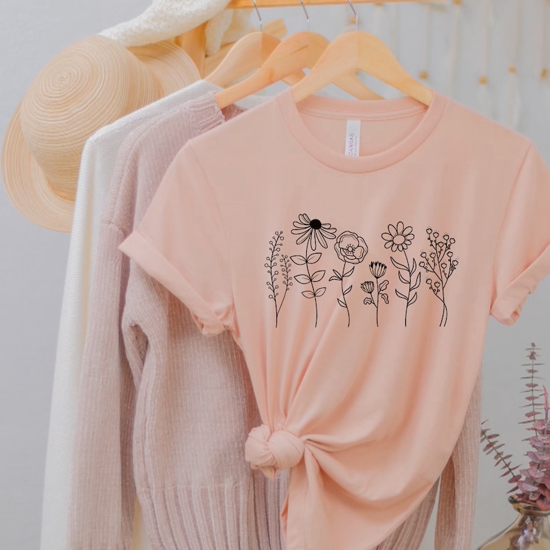 Pu&ograve; includere: Una maglietta rosa pallido con un disegno a linea nera di una fila di fiori e steli.