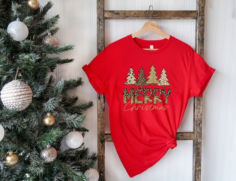 May include: Red t-shirt with a graphic of gold and brown Christmas trees with a leopard print design and the text "Merry Christmas".
