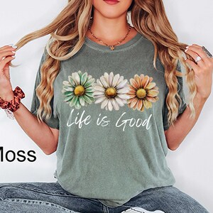May include: Moss green t-shirt featuring three daisy flowers in green, white, and orange. The words "Life is Good" are printed in white script below the flowers. The shirt is a casual style.