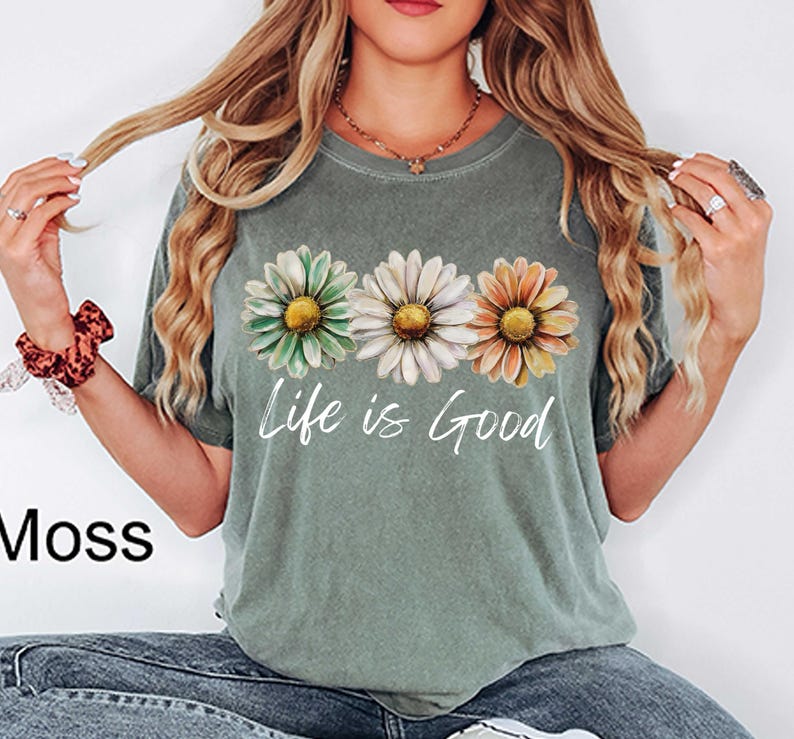 May include: Moss green t-shirt featuring three daisy flowers in green, white, and orange. The words "Life is Good" are printed in white script below the flowers. The shirt is a casual style.