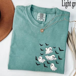 May include: Light green t-shirt with a crew neck and short sleeves. The shirt features a design of white cartoon ghosts and black bats. A silver necklace with a pendant is draped across the shirt.