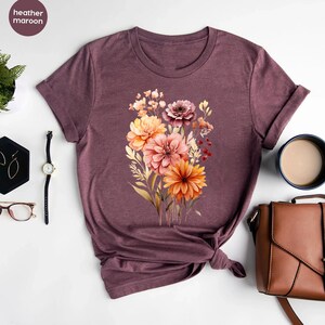 May include: A heather maroon t-shirt with a floral print featuring a bouquet of orange, pink, and brown flowers.