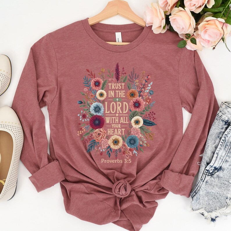 Peut inclure: Un t-shirt &agrave; manches longues de couleur mauve avec un motif floral et le texte "Trust in the Lord with all your heart" et "Proverbes 3:5". Le motif comprend des fleurs et des feuilles color&eacute;es.