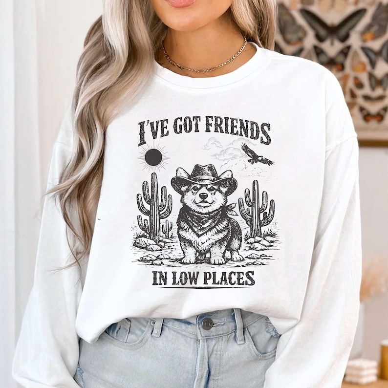 May include: White sweatshirt with a graphic featuring a Corgi wearing a cowboy hat, cacti, and the text "I've Got Friends In Low Places". The design is in black and white, with a vintage, distressed look.