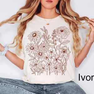 K&ouml;nnte beinhalten: Elfenbeinfarbenes T-Shirt mit einer detaillierten braunen Blumenillustration. Das Design umfasst verschiedene Blumen und Bl&auml;tter. Der Text "Ivory" ist unten rechts auf dem Shirt in Schwarz gedruckt.