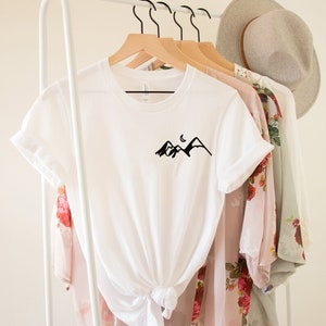 K&ouml;nnte beinhalten: Wei&szlig;es T-Shirt mit einem schwarzen Berg- und Halbmondmotiv auf der Vorderseite. Das Shirt h&auml;ngt an einem wei&szlig;en Kleiderst&auml;nder mit anderer Kleidung.