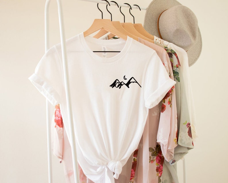 K&ouml;nnte beinhalten: Wei&szlig;es T-Shirt mit einem schwarzen Berg- und Halbmondmotiv auf der Vorderseite. Das Shirt h&auml;ngt an einem wei&szlig;en Kleiderst&auml;nder mit anderer Kleidung.
