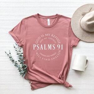 May include: A pink t-shirt with a white circle graphic that says "God is my refuge and my fortress, Psalms 91, I will find shelter under his wings".