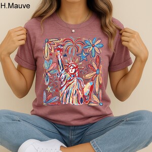 May include: Mauve-colored t-shirt featuring a colorful illustration of the Statue of Liberty, surrounded by flowers, fireworks, and stars. The text "H.Mauve" is in the upper left corner. The shirt has short sleeves and a round neckline.