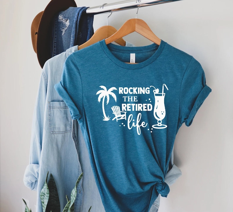 Op de afbeelding: Een tealblauw t-shirt met een wit grafisch ontwerp met de tekst "Rocking the Retired Life". De grafiek bevat ook een palmboom, een strandstoel en een cocktail.