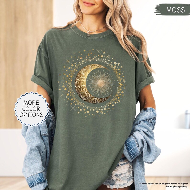 May include: A moss green t-shirt featuring a gold crescent moon and sun design surrounded by stars. The shirt has rolled-up sleeves and the text "MORE COLOR OPTIONS" is visible. The shirt's color may vary slightly due to photography.