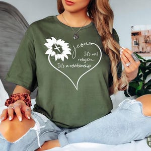 May include: A military green t-shirt featuring a white graphic of a sunflower with a cross in the center, surrounded by a heart outline and the text: Jesus It's not religion... It's a relationship. The shirt has short sleeves and a crew neckline.