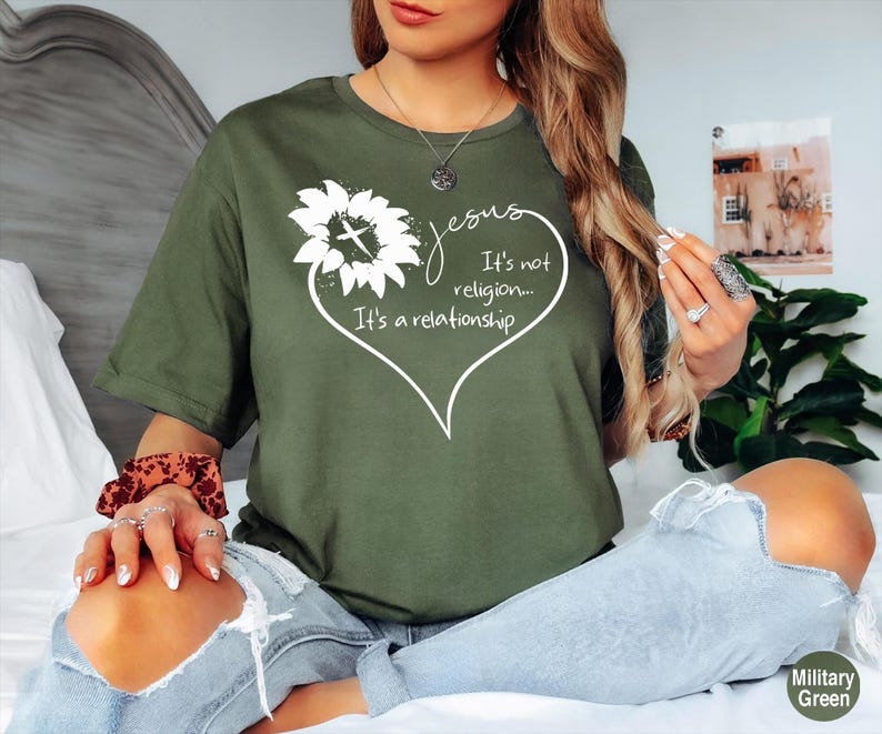 May include: A military green t-shirt featuring a white graphic of a sunflower with a cross in the center, surrounded by a heart outline and the text: Jesus It's not religion... It's a relationship. The shirt has short sleeves and a crew neckline.
