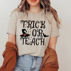 Puede incluir: Camiseta beige con las palabras "TRICK OR TEACH" en escritura negra, con un sombrero de bruja y un gr&aacute;fico de murci&eacute;lago. La camiseta es de estilo cuello en V y la lleva una persona.