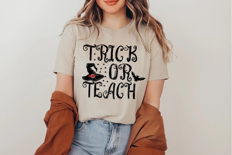 Puede incluir: Camiseta beige con las palabras "TRICK OR TEACH" en escritura negra, con un sombrero de bruja y un gr&aacute;fico de murci&eacute;lago. La camiseta es de estilo cuello en V y la lleva una persona.