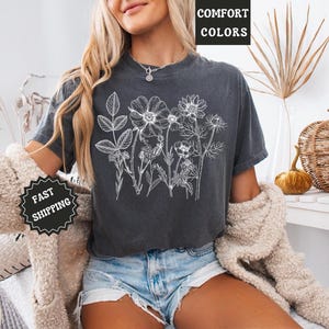 May include: Dark gray t-shirt featuring a white floral design. The shirt has the words "COMFORT COLORS" printed at the top. The model is wearing denim shorts and a cream-colored sweater. A black and white "FAST SHIPPING" sticker is visible.