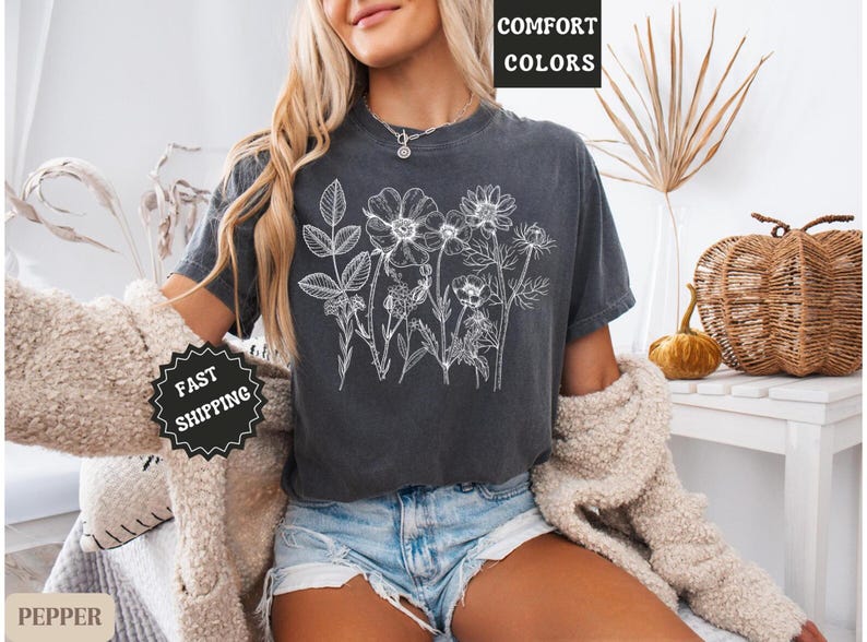 May include: Dark gray t-shirt featuring a white floral design. The shirt has the words "COMFORT COLORS" printed at the top. The model is wearing denim shorts and a cream-colored sweater. A black and white "FAST SHIPPING" sticker is visible.