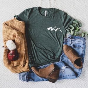K&ouml;nnte beinhalten: Ein gr&uuml;nes T-Shirt mit einem wei&szlig;en Berg- und Halbmondmotiv. Das T-Shirt liegt auf einer wei&szlig;en und grauen Chevron-Oberfl&auml;che mit einem braunen Pullover, blauen Jeans und braunen Stiefeletten.