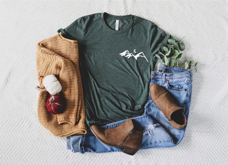 K&ouml;nnte beinhalten: Ein gr&uuml;nes T-Shirt mit einem wei&szlig;en Berg- und Halbmondmotiv. Das T-Shirt liegt auf einer wei&szlig;en und grauen Chevron-Oberfl&auml;che mit einem braunen Pullover, blauen Jeans und braunen Stiefeletten.