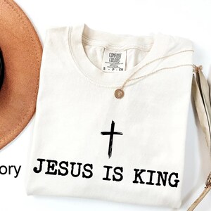 K&ouml;nnte beinhalten: Elfenbeinfarbenes T-Shirt mit Rundhalsausschnitt, mit einem schwarzen Kreuz und dem Text "JESUS IS KING". Eine goldene Halskette mit Anh&auml;nger liegt auf dem Shirt. Ein brauner Hut und eine getrocknete Pflanze sind im Hintergrund.