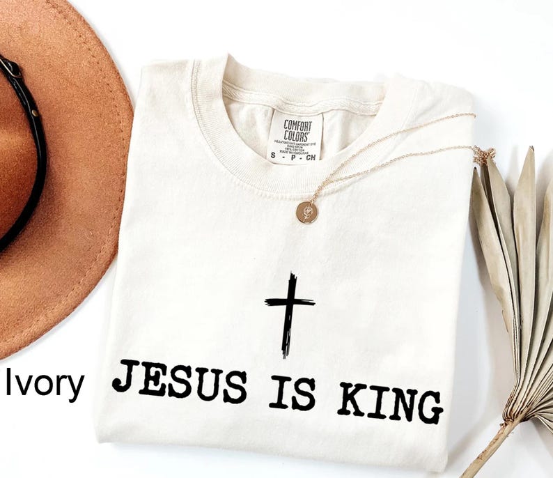 K&ouml;nnte beinhalten: Elfenbeinfarbenes T-Shirt mit Rundhalsausschnitt, mit einem schwarzen Kreuz und dem Text "JESUS IS KING". Eine goldene Halskette mit Anh&auml;nger liegt auf dem Shirt. Ein brauner Hut und eine getrocknete Pflanze sind im Hintergrund.