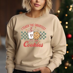 May include: A tan sweatshirt with a retro-style graphic of a glass of milk and a cookie. The text on the sweatshirt reads "I teach the smartest cookies".
