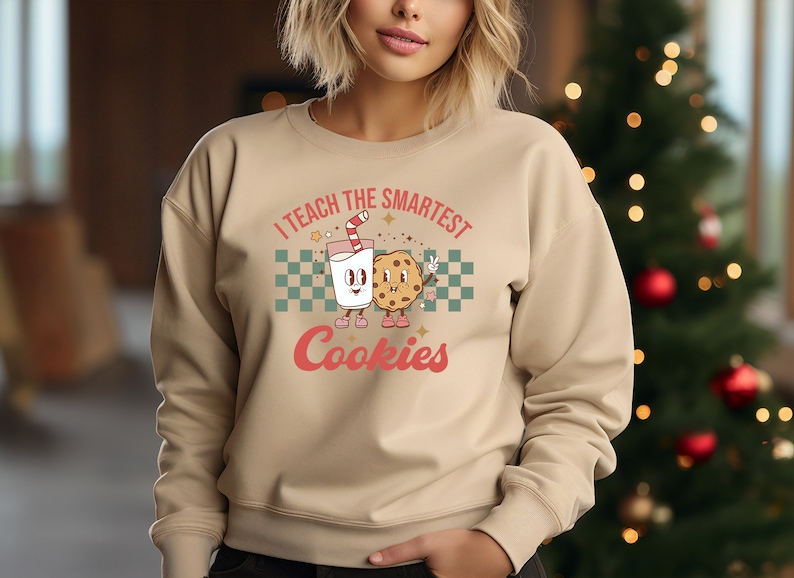 May include: A tan sweatshirt with a retro-style graphic of a glass of milk and a cookie. The text on the sweatshirt reads "I teach the smartest cookies".