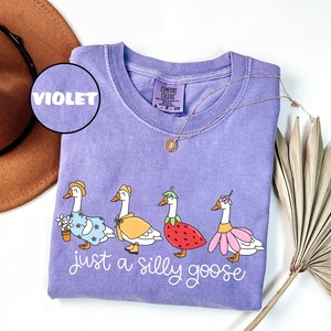 May include: A violet Comfort Colors t-shirt featuring a whimsical design of four geese dressed in various outfits. The phrase "just a silly goose" is written in cursive below the geese. A gold necklace and a brown hat are also visible.