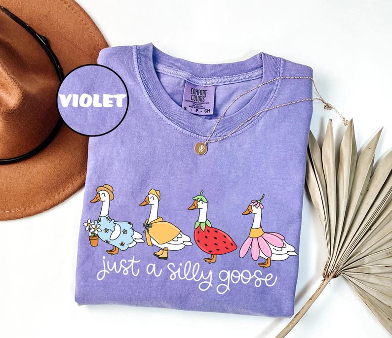 May include: A violet Comfort Colors t-shirt featuring a whimsical design of four geese dressed in various outfits. The phrase "just a silly goose" is written in cursive below the geese. A gold necklace and a brown hat are also visible.