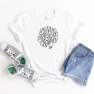 May include: White t-shirt with black text that says "Stay Groovy Baby". The shirt is paired with denim shorts and gray sneakers.