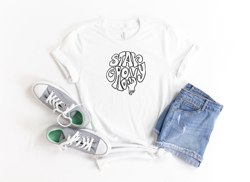 May include: White t-shirt with black text that says "Stay Groovy Baby". The shirt is paired with denim shorts and gray sneakers.