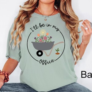 Op de afbeelding: Een lichtgroen T-shirt met de tekst "I'll be in my office" in zwarte letters. De tekst wordt omringd door een cirkel met een kleine kruiwagen vol kleurrijke bloemen.