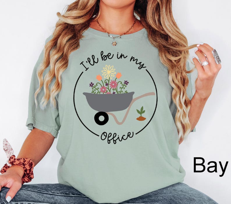 Op de afbeelding: Een lichtgroen T-shirt met de tekst "I'll be in my office" in zwarte letters. De tekst wordt omringd door een cirkel met een kleine kruiwagen vol kleurrijke bloemen.