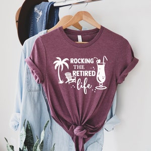 Op de afbeelding: Een maroon t-shirt met witte tekst die "Rocking the Retired life." luidt. Het shirt heeft een palmboom, een strandstoel en een cocktailglas met een kers en een rietje.