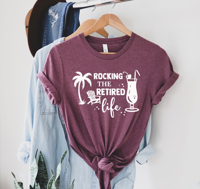 Op de afbeelding: Een maroon t-shirt met witte tekst die "Rocking the Retired life." luidt. Het shirt heeft een palmboom, een strandstoel en een cocktailglas met een kers en een rietje.