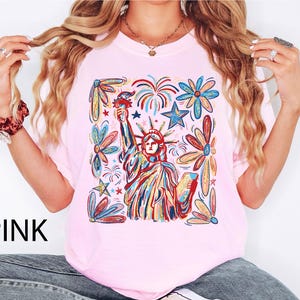 May include: Pink t-shirt featuring a colorful illustration of the Statue of Liberty holding a torch, surrounded by fireworks and floral designs. The word "PINK" is printed in black at the bottom left.
