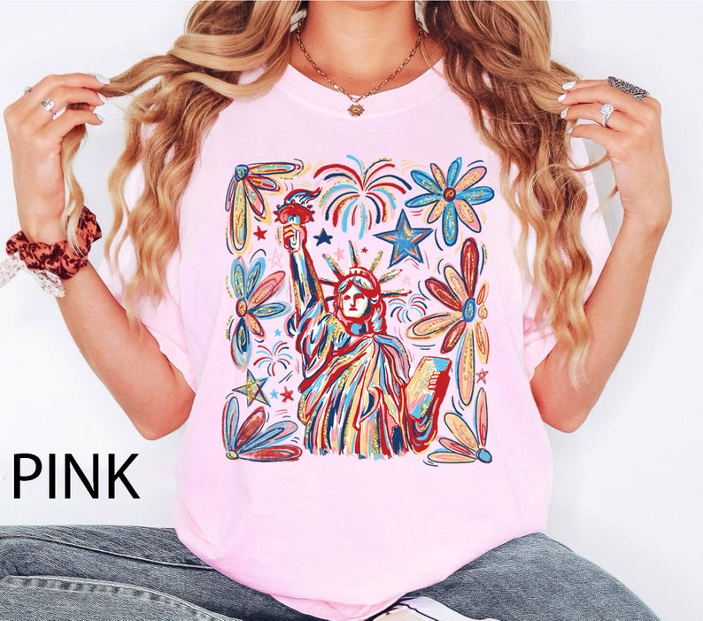 May include: Pink t-shirt featuring a colorful illustration of the Statue of Liberty holding a torch, surrounded by fireworks and floral designs. The word "PINK" is printed in black at the bottom left.