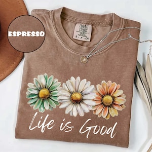 May include: Espresso-colored t-shirt with a daisy graphic in shades of green, white, and orange. The words "Life is Good" are printed in white script below the flowers. A silver necklace with a pendant is draped across the shirt.