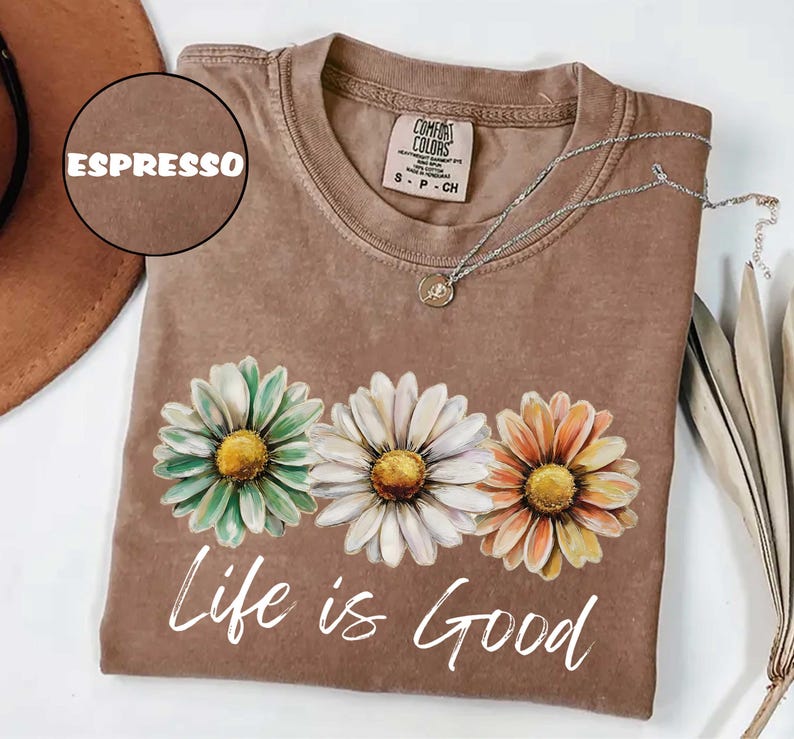 May include: Espresso-colored t-shirt with a daisy graphic in shades of green, white, and orange. The words "Life is Good" are printed in white script below the flowers. A silver necklace with a pendant is draped across the shirt.