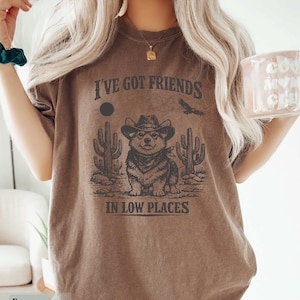 May include: Brown t-shirt with a graphic of a corgi wearing a cowboy hat and bandana, with cacti and the text "I've Got Friends In Low Places". A sun and bird are also in the design. The shirt is a neutral color.